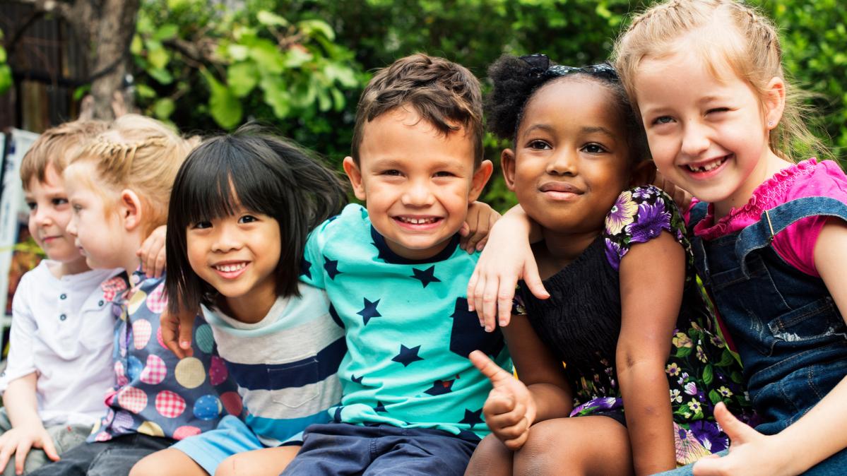 A group of kindergarten aged children sit in a row outdoors with their arms around each other smiling.